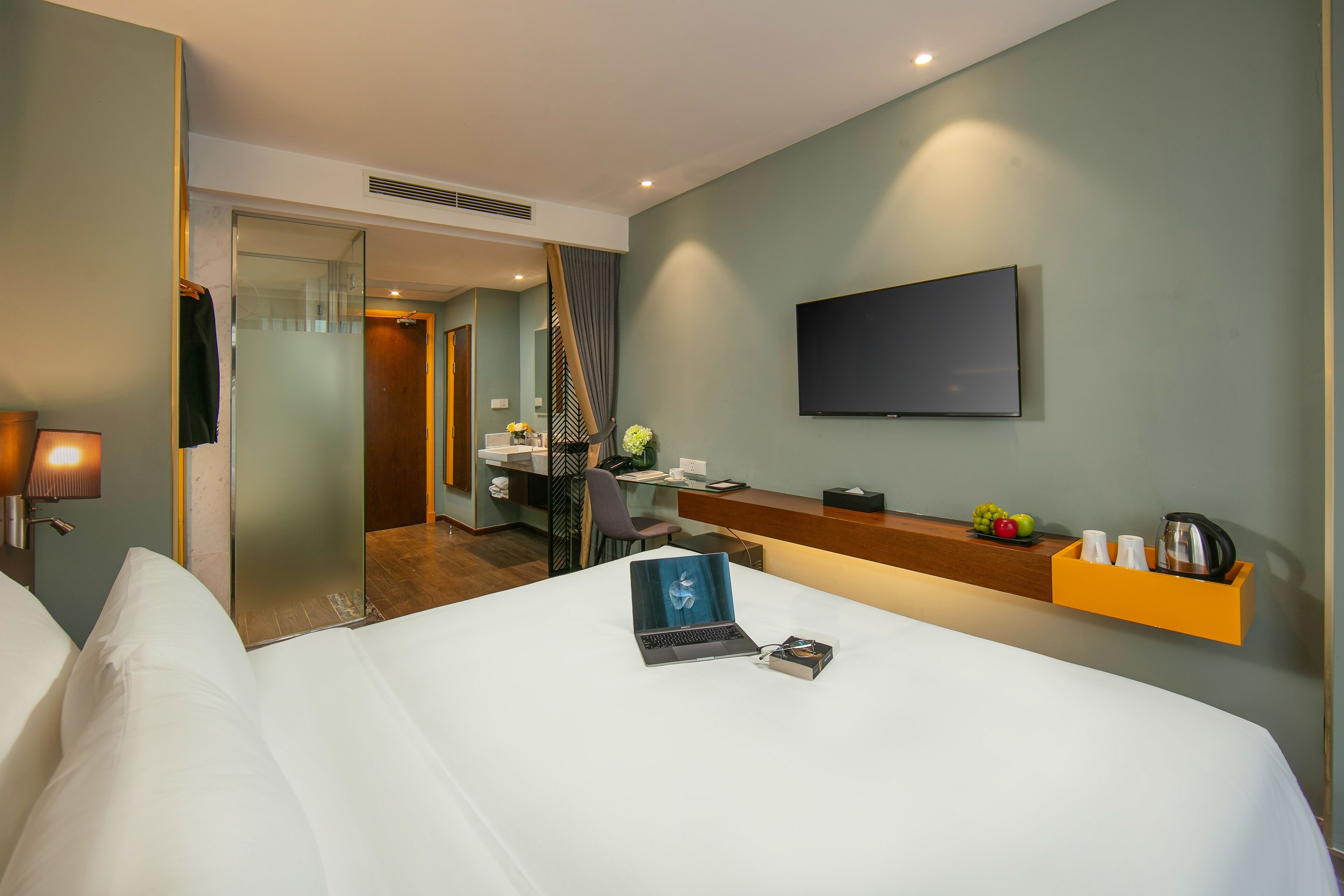 Habitación de hotel con cama, escritorio, silla, televisión y una pequeña estantería con frutas y tetera.