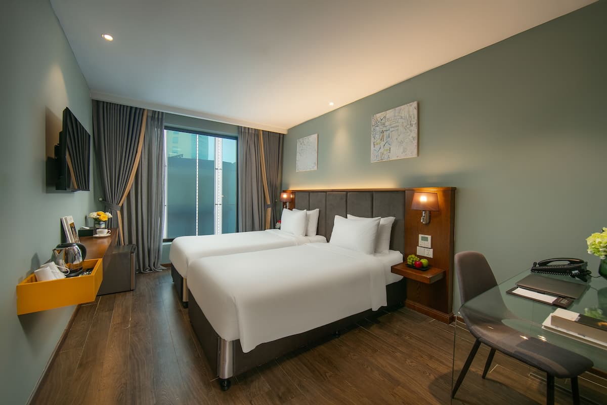 Ett hotellrum med två sängar, ett skrivbord med en stol, en tv som är monterad på väggen och ett fönster med gardiner.