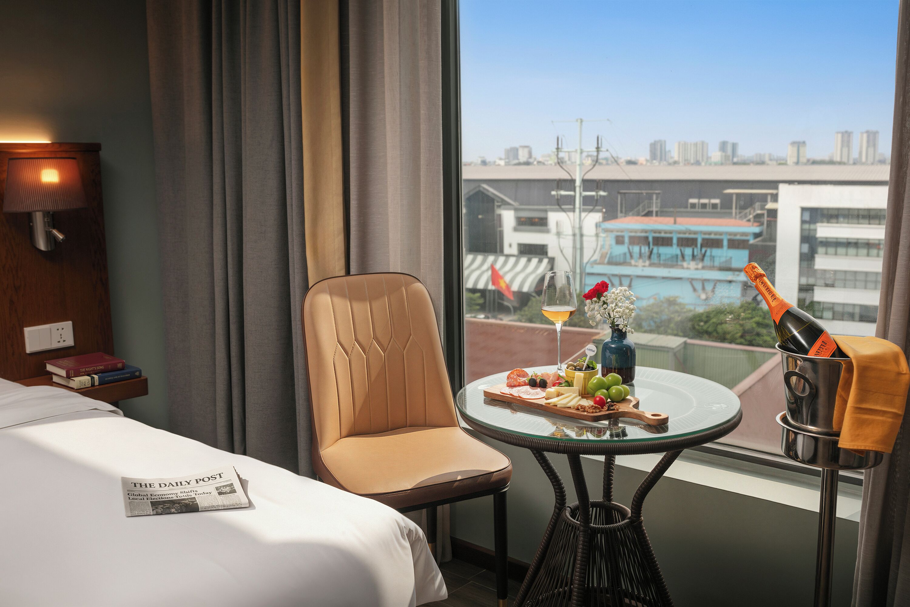 Une chambre d’hôtel avec un lit, une chaise, une petite table sur laquelle se trouve une bouteille de champagne, un plateau avec de la nourriture et un journal.