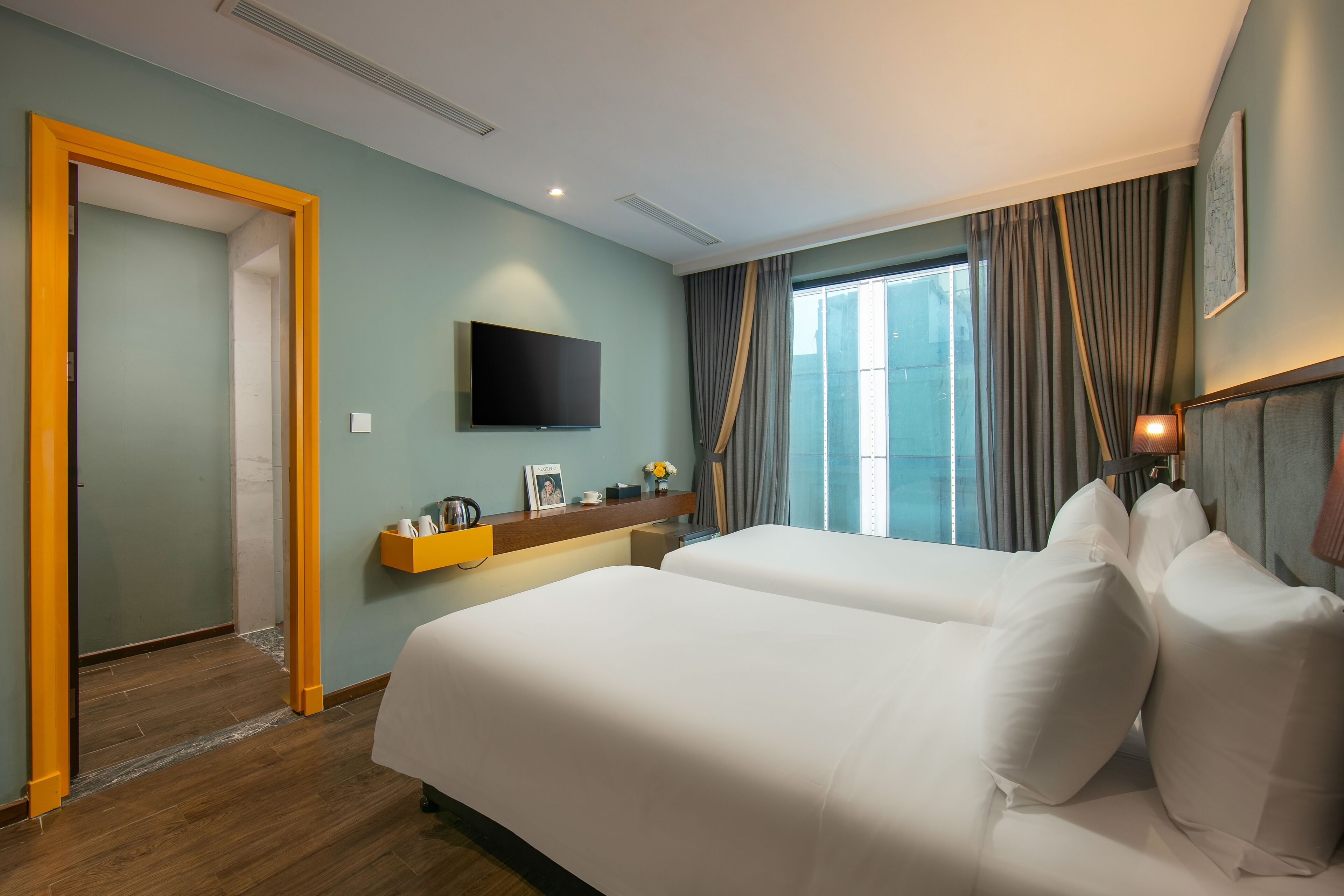 A hotel room with two beds, a TV mounted on the wall, a window with curtains, and a wooden floor.