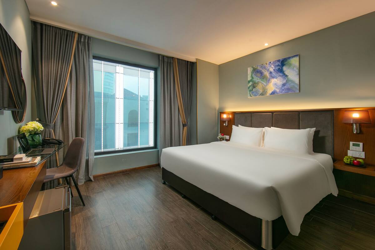 Ett modernt hotellrum med en stor säng, ett skrivbord med en bärbar dator, en stol och utsikt över staden genom ett stort fönster.