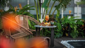 Wicker chairs, a small round table with a drink, fruit, and a bowl, surrounded by lush green plants. - Grand Cititel Hanoi Hotel & Spa (Hanoi)