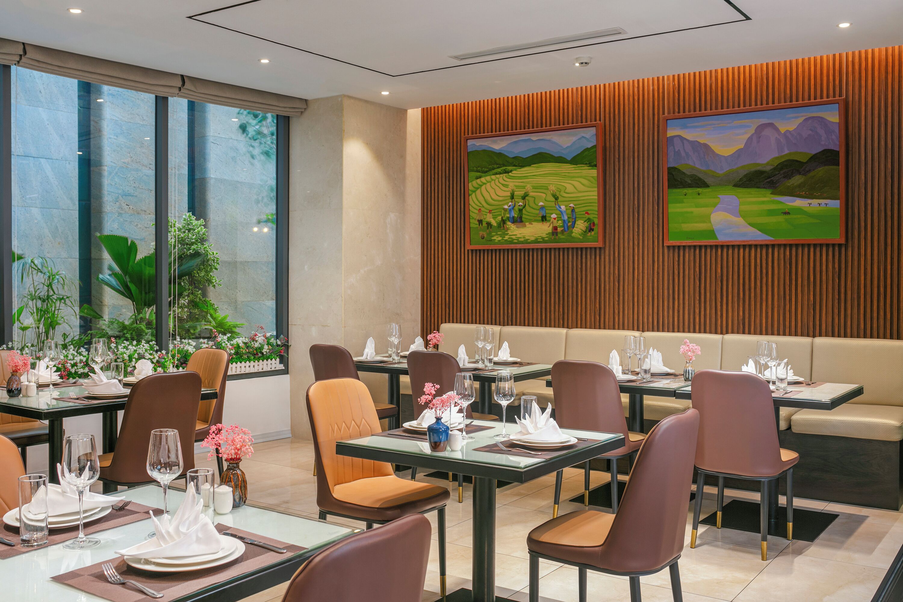 A restaurant with tables set for dining, large windows, and two paintings on the wall.