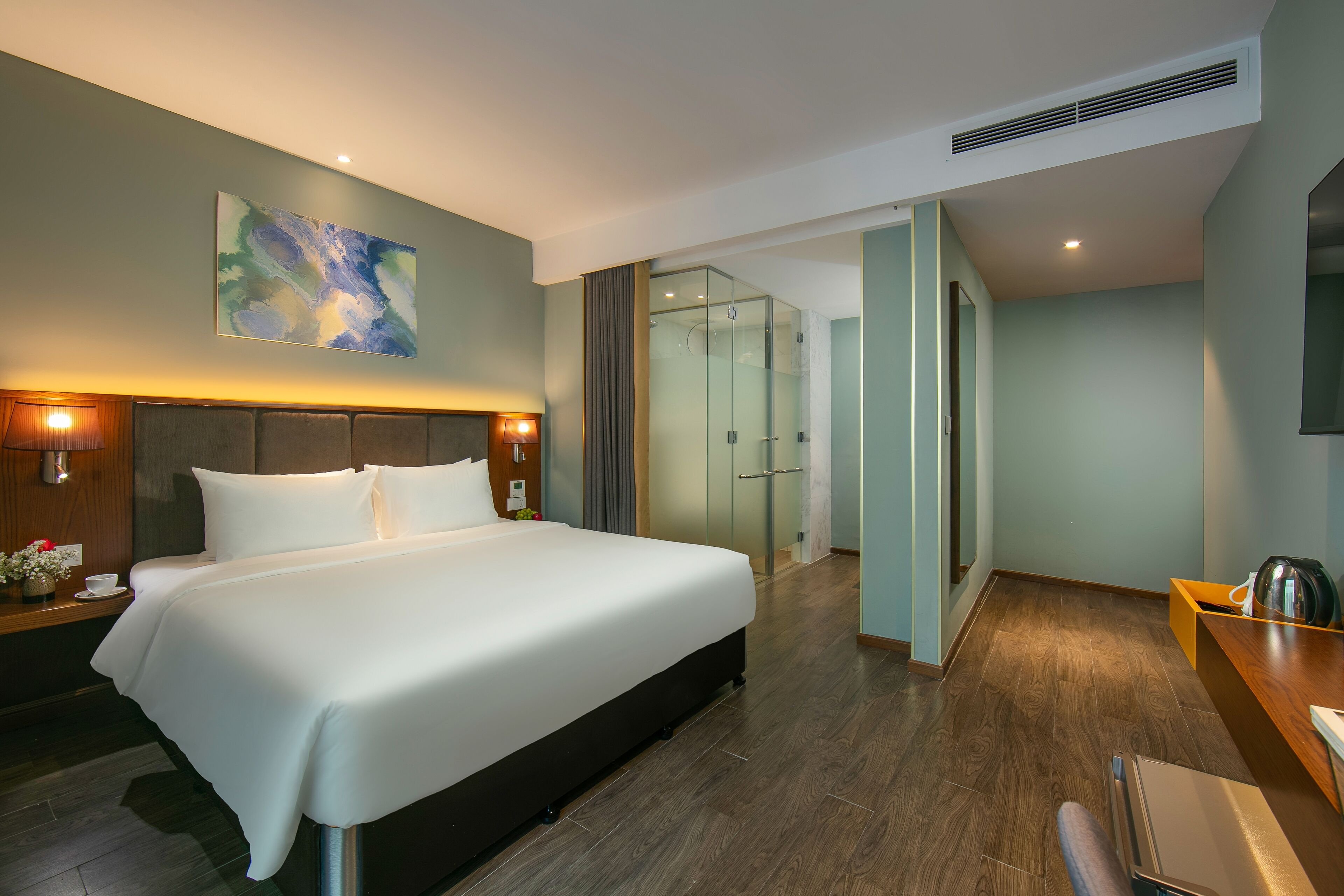 A modern hotel room with a large bed, a desk, a glass shower, and a painting on the wall.