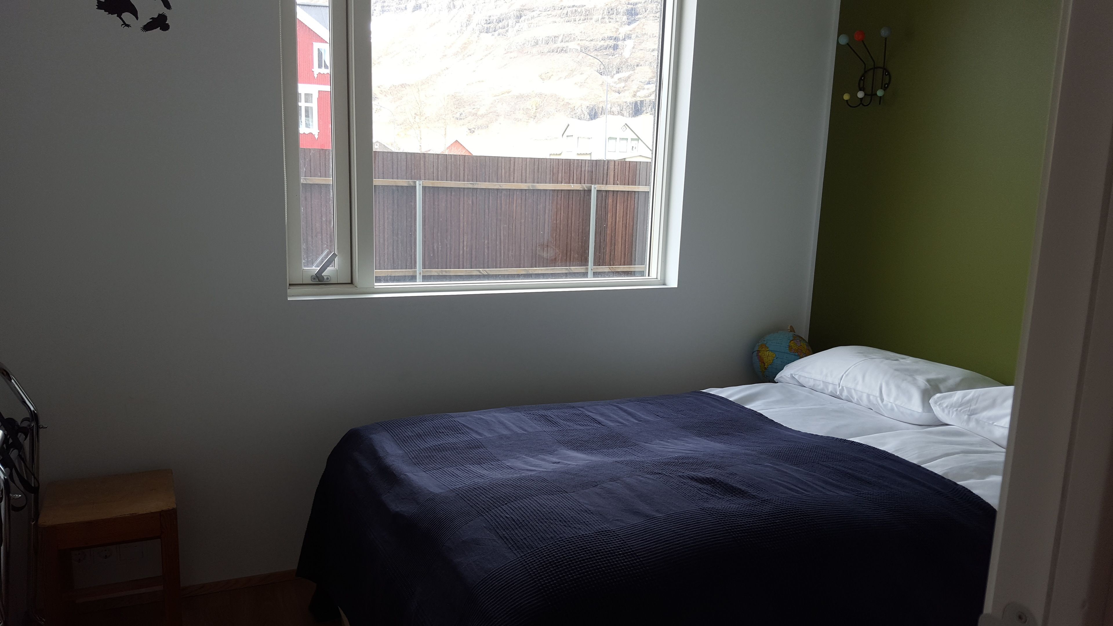 A bedroom with a bed, a small stool, a window, and a view of a red building outside.