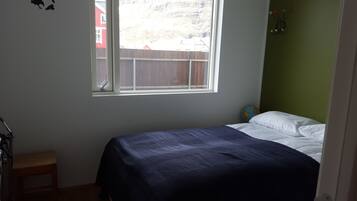 A bedroom with a bed, a small stool, a window, and a view of a red building outside.