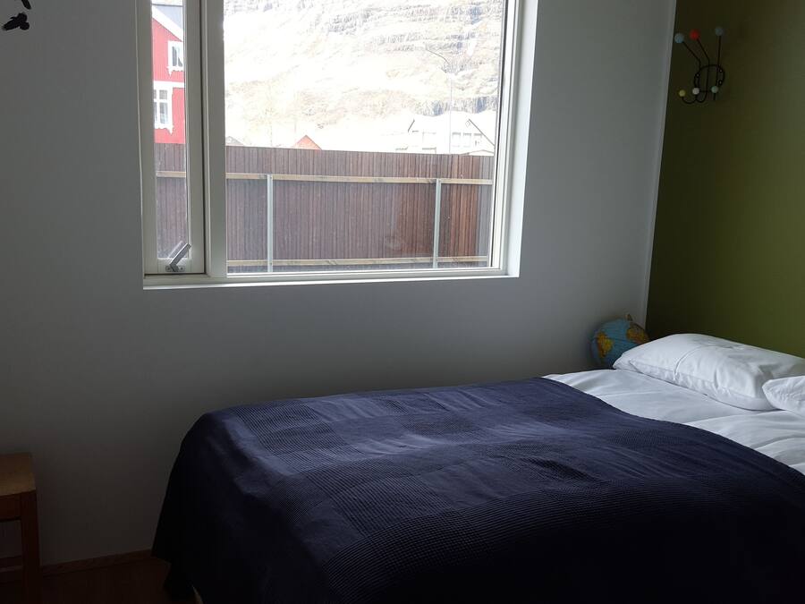 A bedroom with a bed, a small stool, a window, and a view of a red building outside.