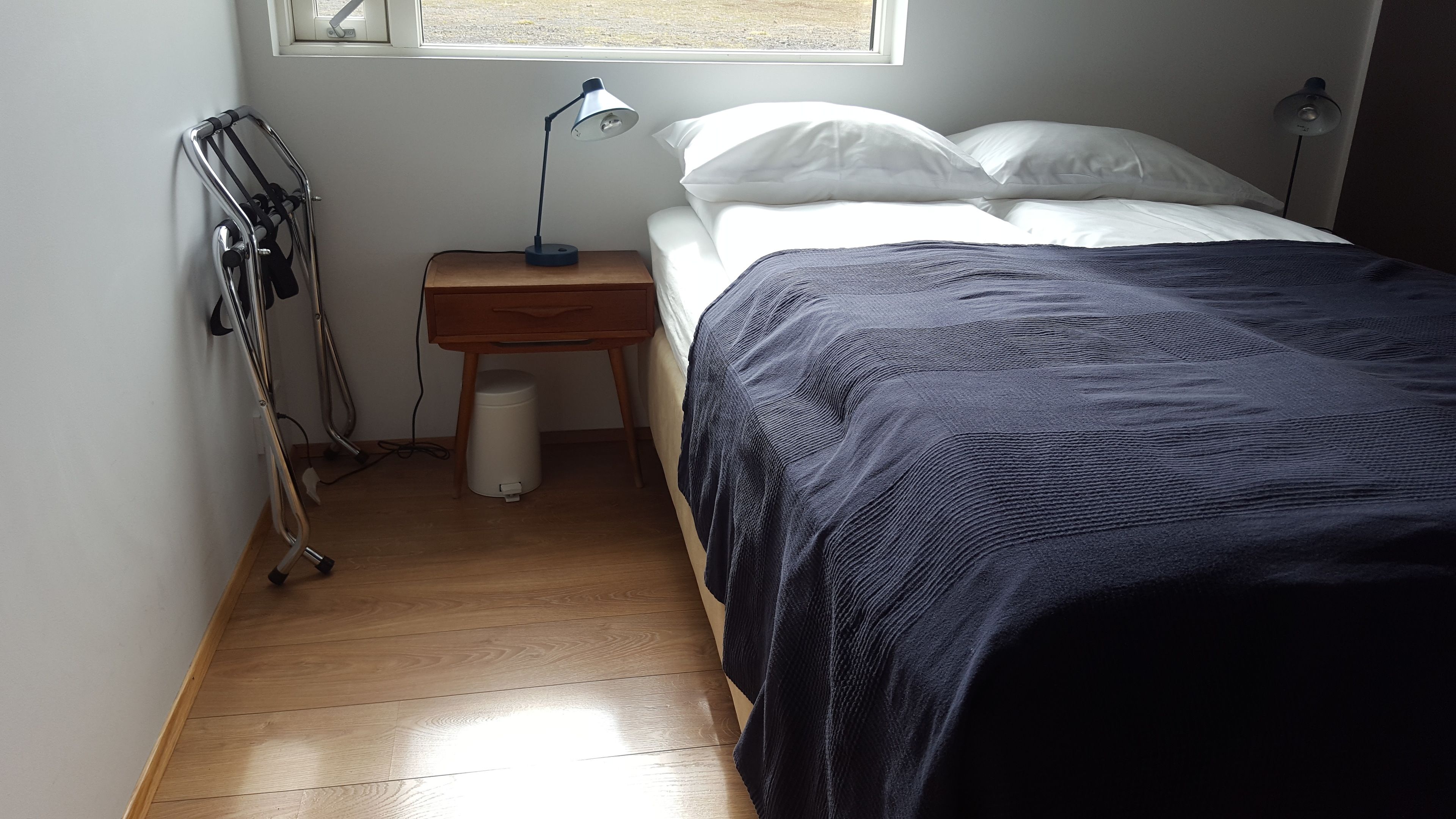 A bedroom with a bed, a nightstand, a lamp, a trash bin, and a window.