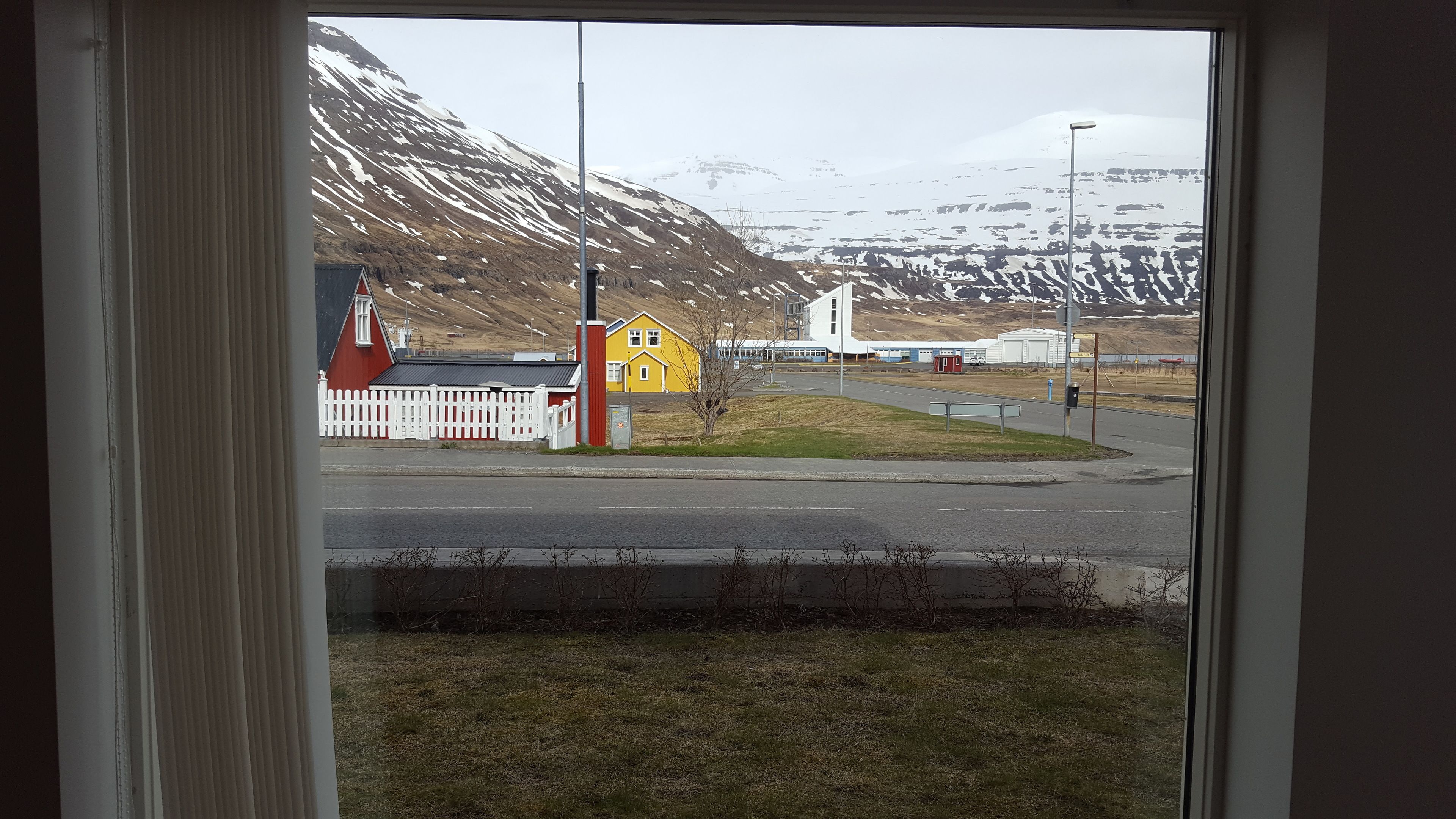 Blick durch ein Fenster auf ein Gebäude, eine Straße, ein kleines gelbes Haus und einen schneebedeckten Berg.