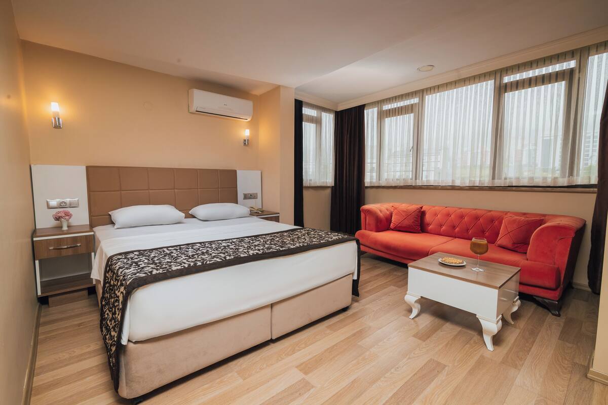 A hotel room with a bed, a red sofa, a small white table, and a window with curtains.