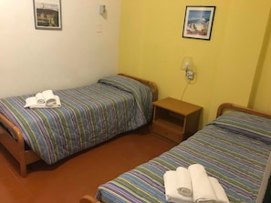 A small, simple room with two single beds, each with a striped bedspread and rolled towels. There is a small wooden nightstand between the beds, and a wall socket above it.