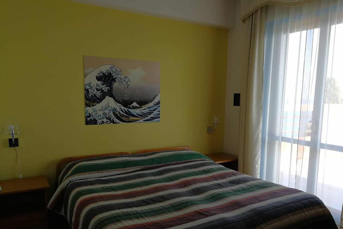 A hotel room with a bed, a painting of a wave on the wall, and a window with curtains.