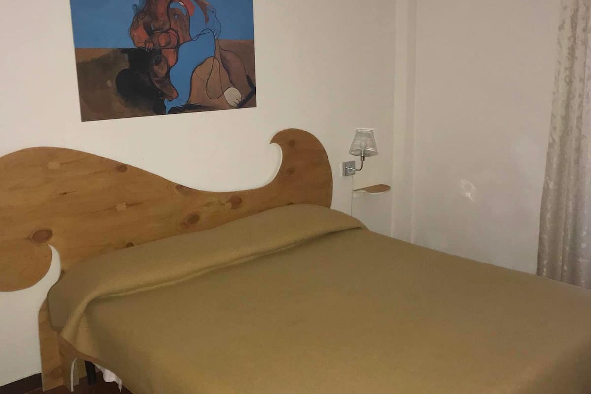 A bedroom with a wooden bed, a beige bedspread, a wall-mounted air conditioner, a small bedside table with a lamp, and a painting of a person on the wall.