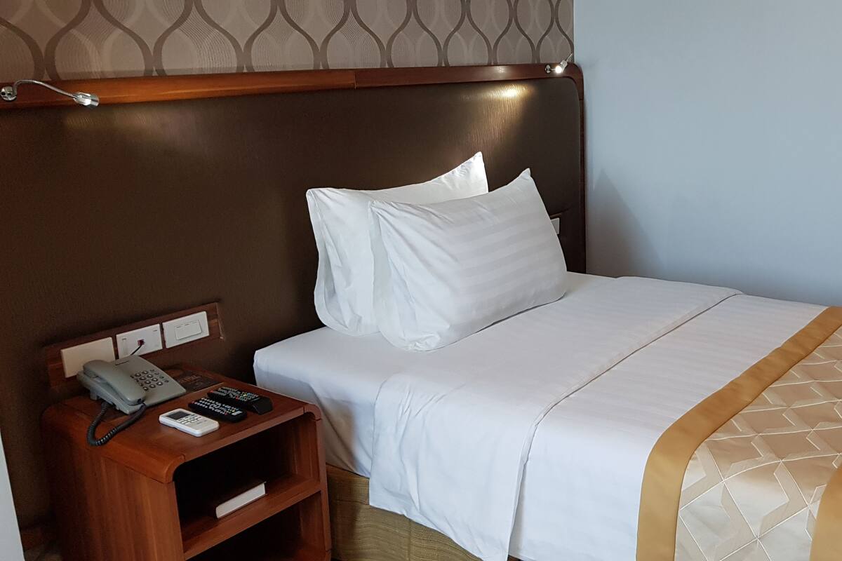 Une chambre d’hôtel avec un lit, une table de chevet en bois sur laquelle se trouvent un téléphone et des télécommandes, et une tête de lit à motifs.