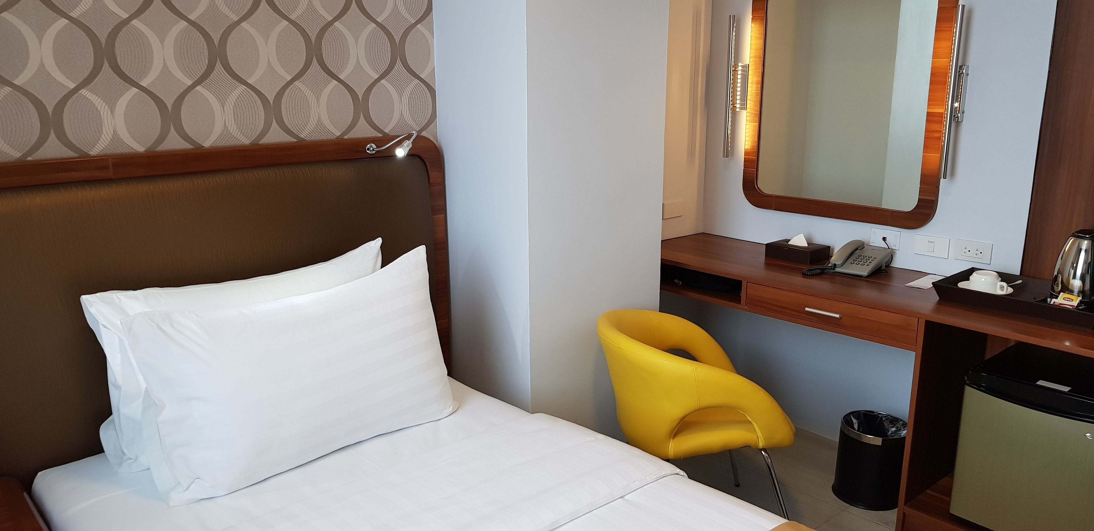 A hotel room with a bed, a desk, a chair, a mirror, a telephone, and a trash bin.