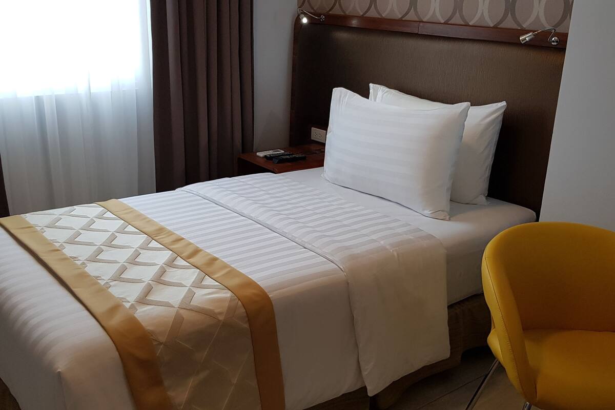 Une chambre d’hôtel avec un grand lit, une tête de lit, une table de chevet, une chaise jaune et une fenêtre avec des rideaux.