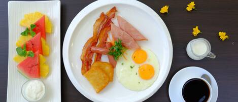 Un plato de desayuno con huevos, tocino, jamón y tostadas, acompañado de fruta y un tazón pequeño de yogur.