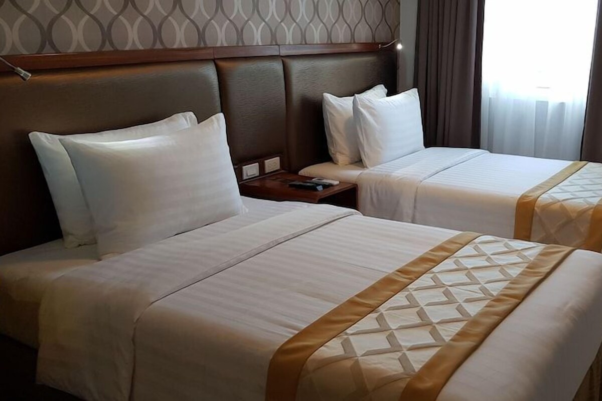 A hotel room with two beds, each with white linens and a gold accent. There is a wooden headboard, a nightstand, and a chair.