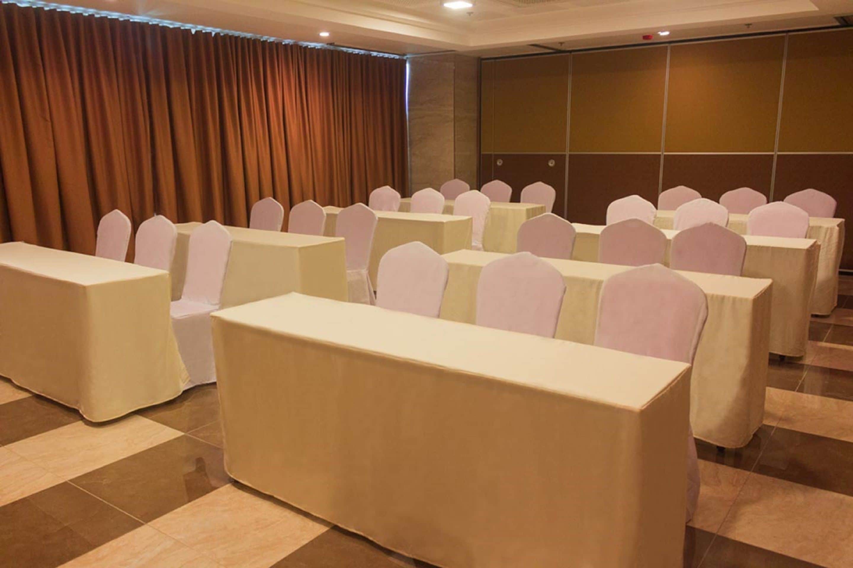 Une salle de conférence avec des rangées de tables et de chaises, un grand écran et un rideau en fond.