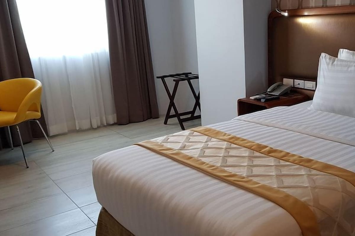 Quarto de hotel com uma cama grande, mesa de cabeceira com telefone, uma cadeira e janela com cortinas.