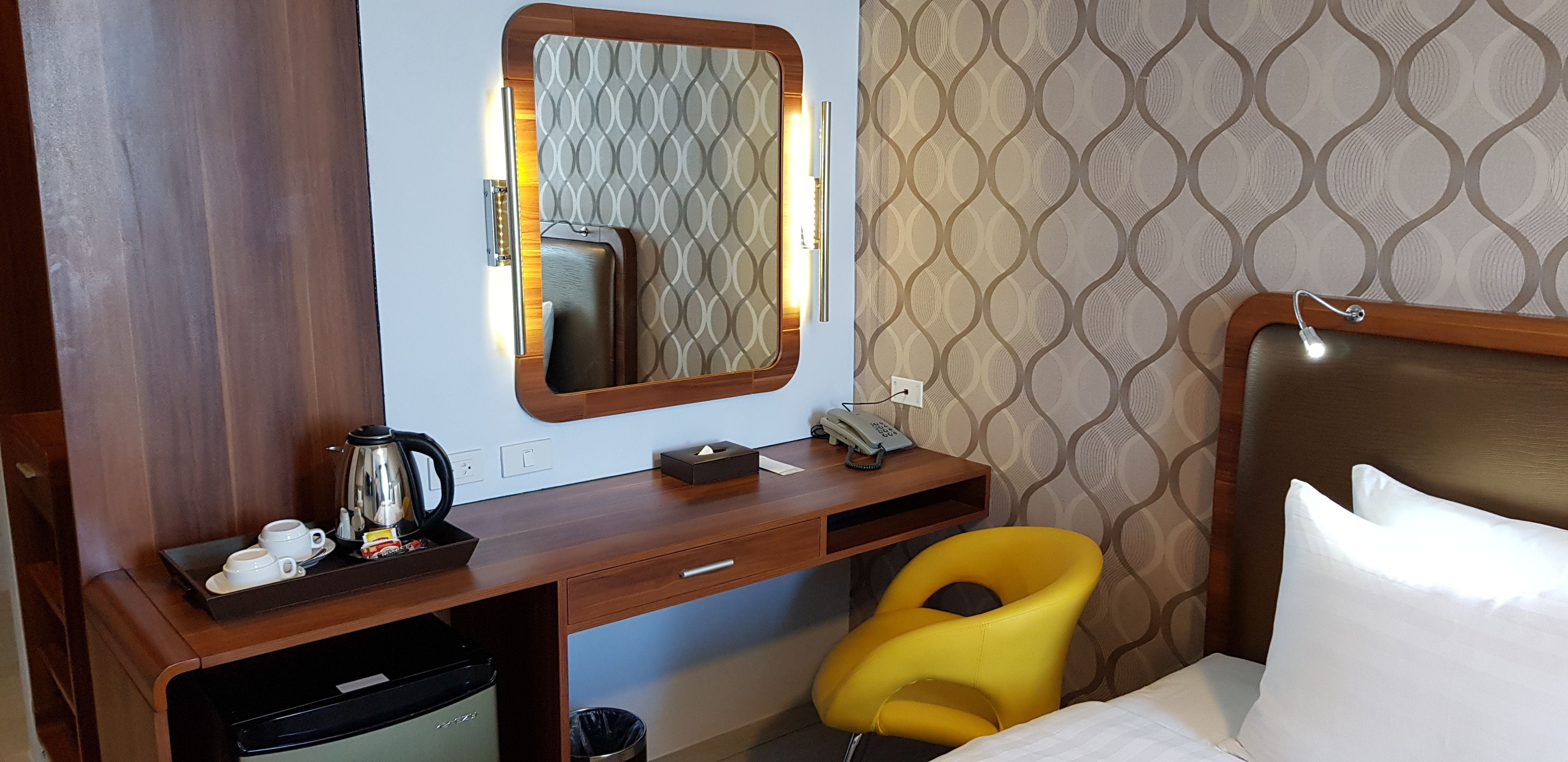 Quarto de hotel pequeno com escrivaninha de madeira, uma cadeira amarela, cama com travesseiros brancos, telefone ao lado da cama e uma pequena lixeira.