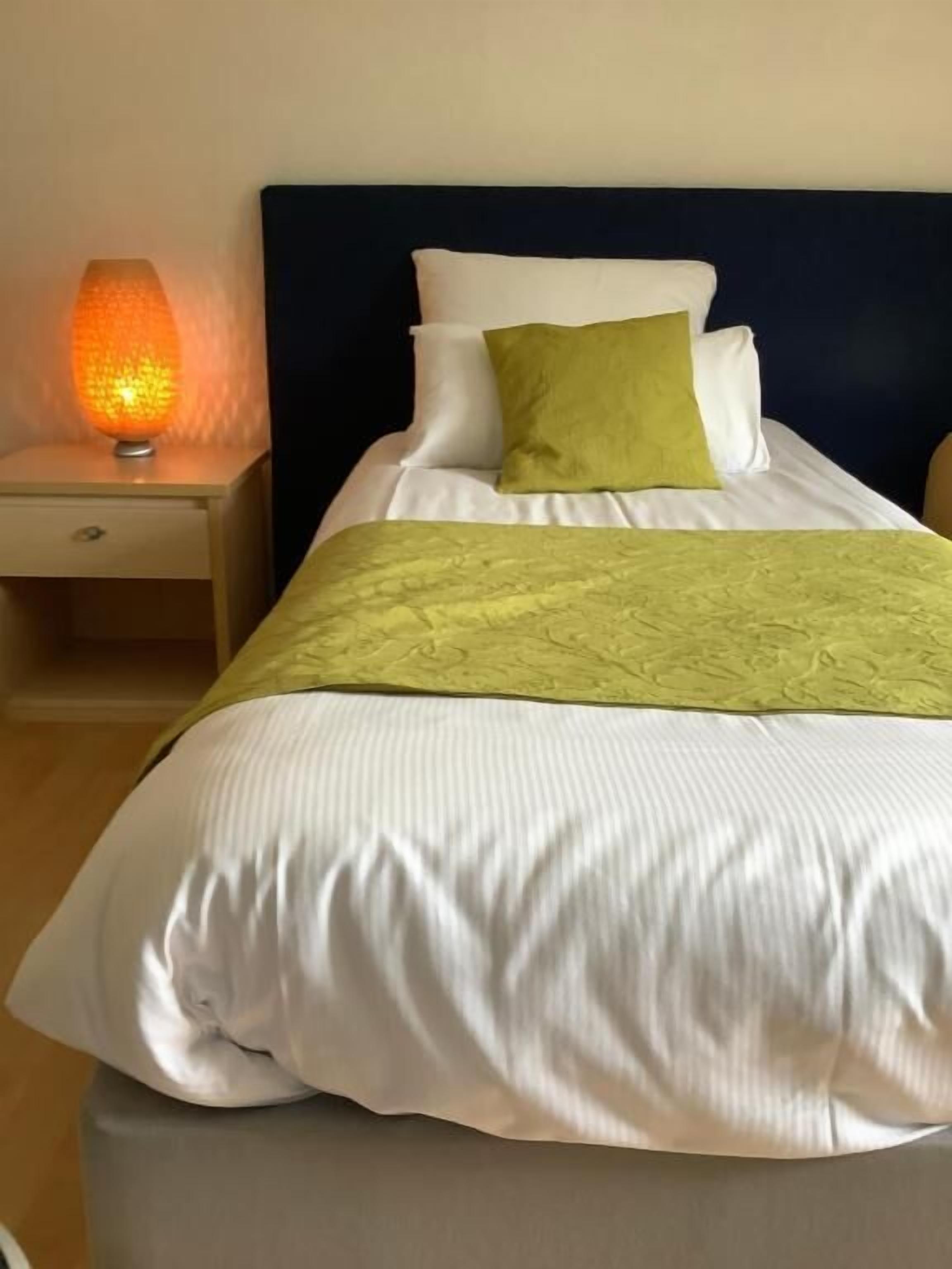 A neatly made bed with white bedding and a green throw, a bedside lamp, and a dark headboard.