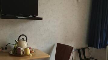 A hotel room with a bed, a table with a kettle and teapot, a chair, a TV mounted on the wall, and a window with curtains.