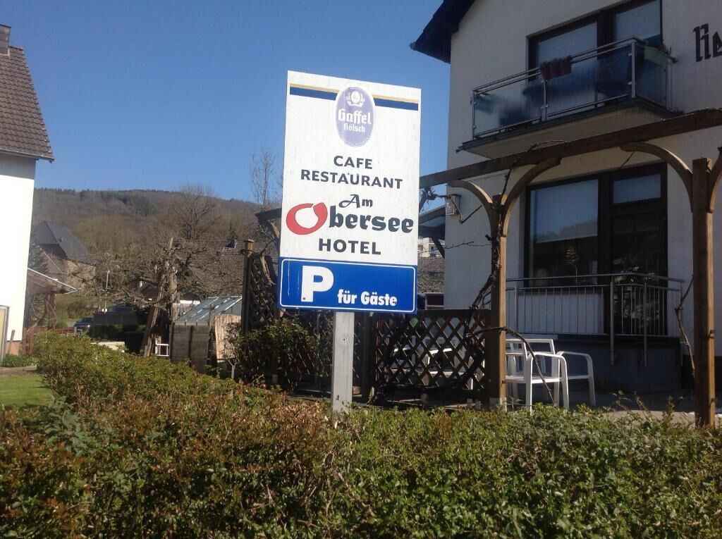 A sign for a cafe, restaurant, and hotel named 'Am Obersee' with a parking sign for guests.