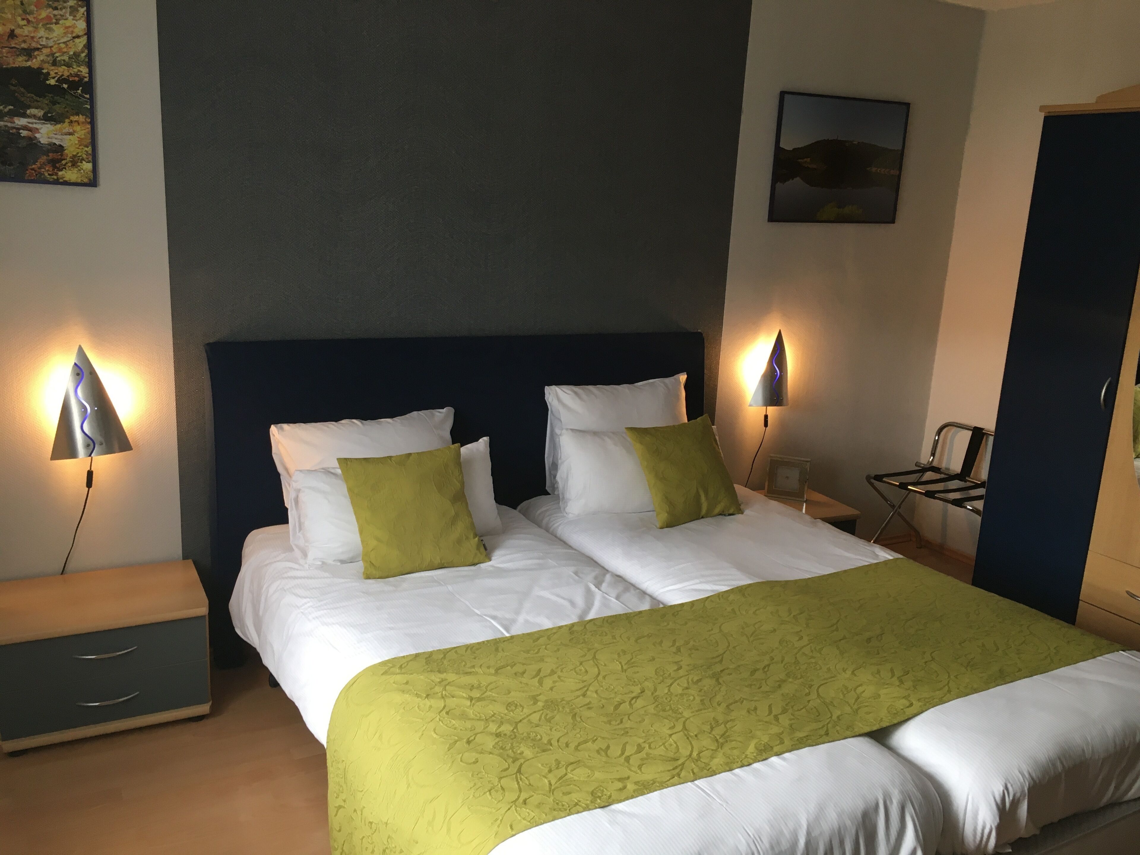Una cama doble con ropa de cama blanca y verde, dos lámparas de noche, una mesita de noche, una silla y un cuadro en la pared.