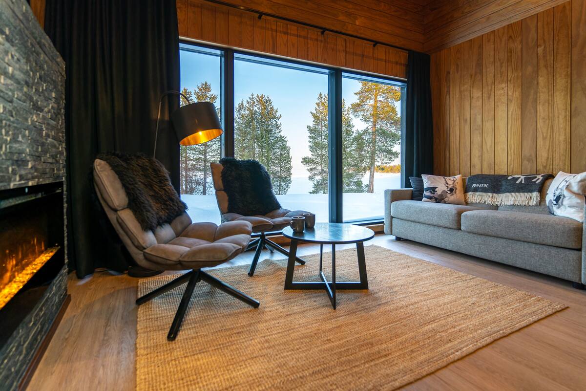 A modern living room with a fireplace, leather armchair, sofa, and large windows offering a view of trees.