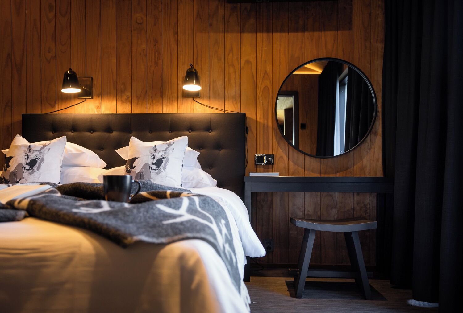 A modern bedroom with a tufted headboard, a round mirror, a bedside table, and a bed with patterned bedding.