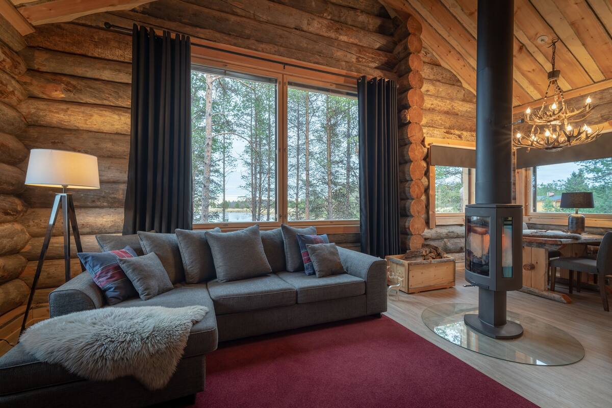 A log cabin interior with a sofa, a lamp, a fireplace, and a view of the forest.