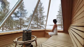 A person sitting in a wooden cabin with a large window, a wood-burning stove, and a view of a forest.