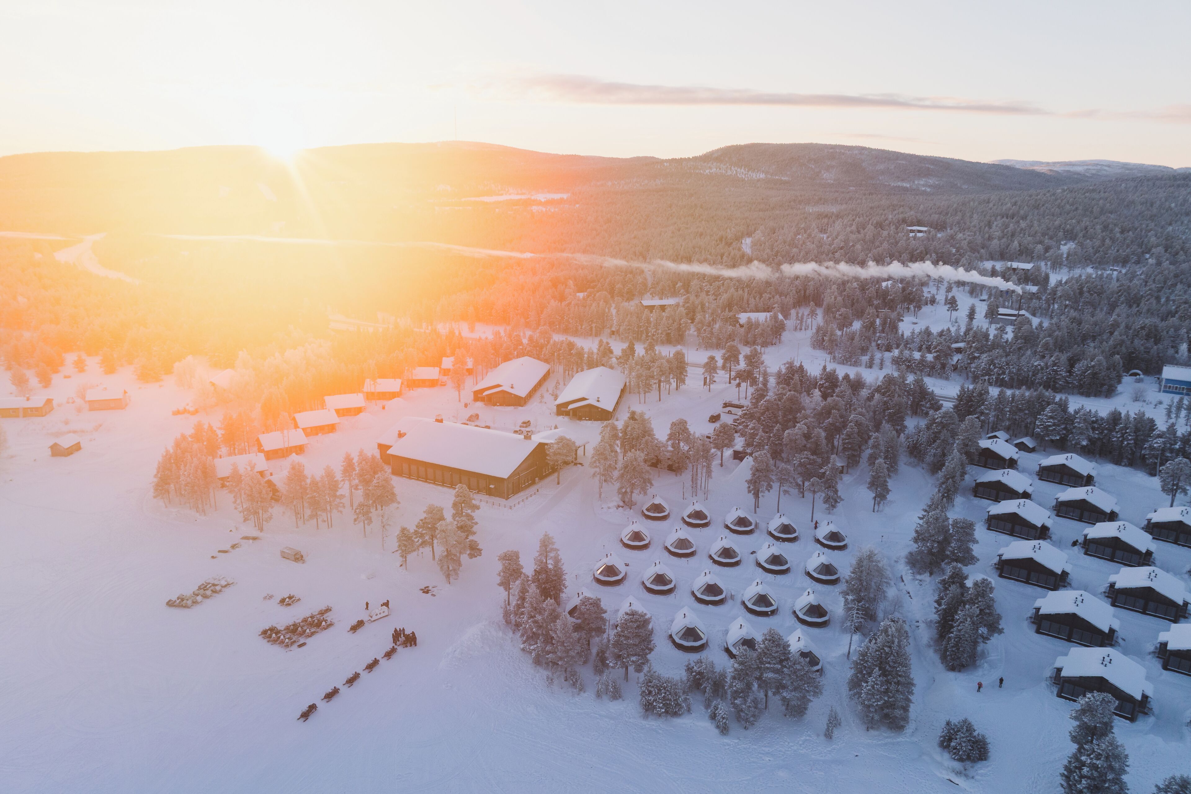 An aerial view of a snow-covered resort with multiple cabins and a central building, surrounded by dense forest.