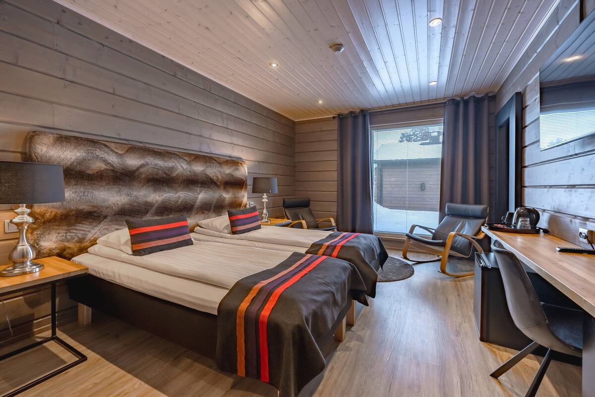 A modern hotel room with a wooden ceiling, two beds with striped bedding, a desk, and a chair.