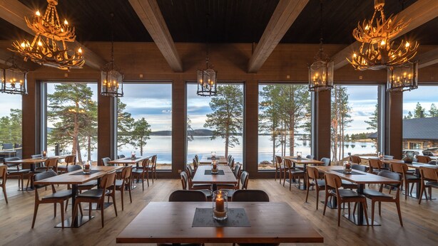 A restaurant with large windows offering a view of a lake and trees. Tables are arranged for dining, and there are multiple chandeliers hanging from the ceiling. - Wilderness Hotel Inari & Igloos (Inari)