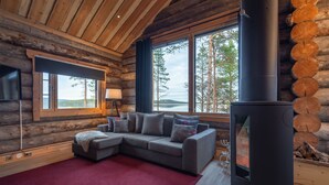 A log cabin interior with a sofa, a fireplace, and a large window overlooking a lake. - Wilderness Hotel Inari & Igloos (Inari)