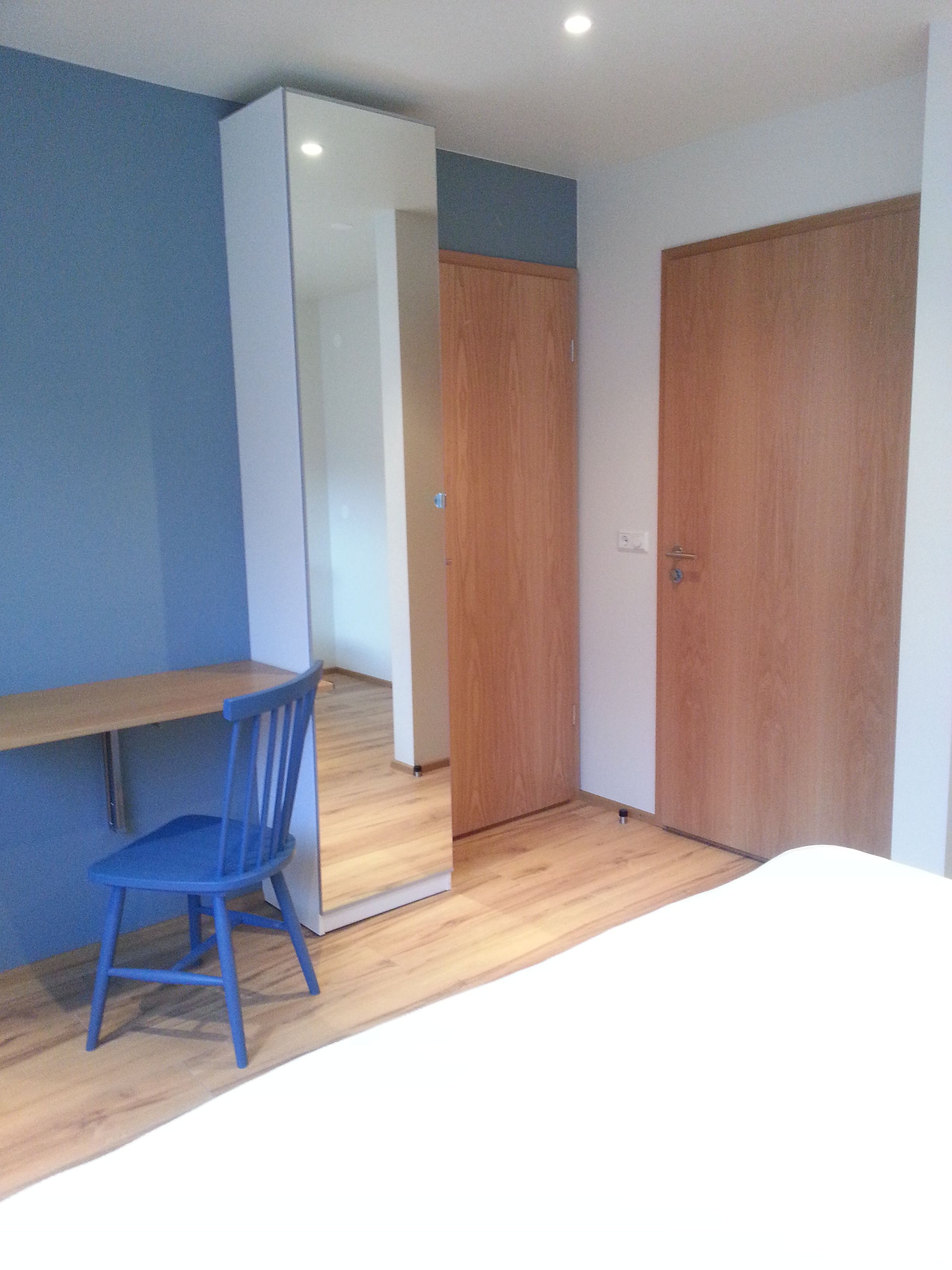 A room with a wooden desk, a blue chair, a white wardrobe, and a wooden door.