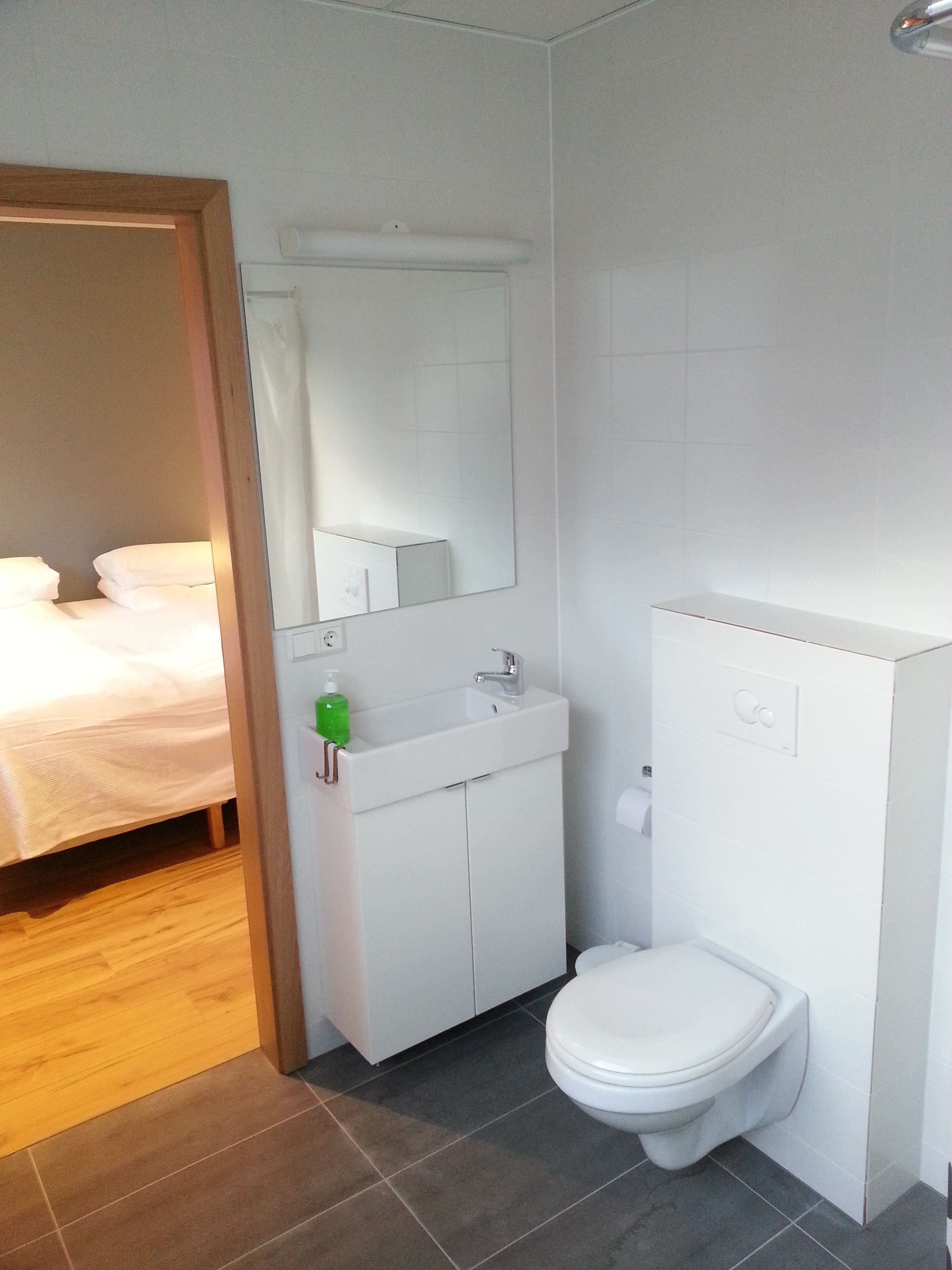 Un baño moderno con lavamanos, espejo y retrete blancos. El suelo es de madera y se ve una cama en la habitación contigua.