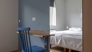 A room with a wooden floor, a blue chair, a desk, a bed with white bedding, and a window with curtains.