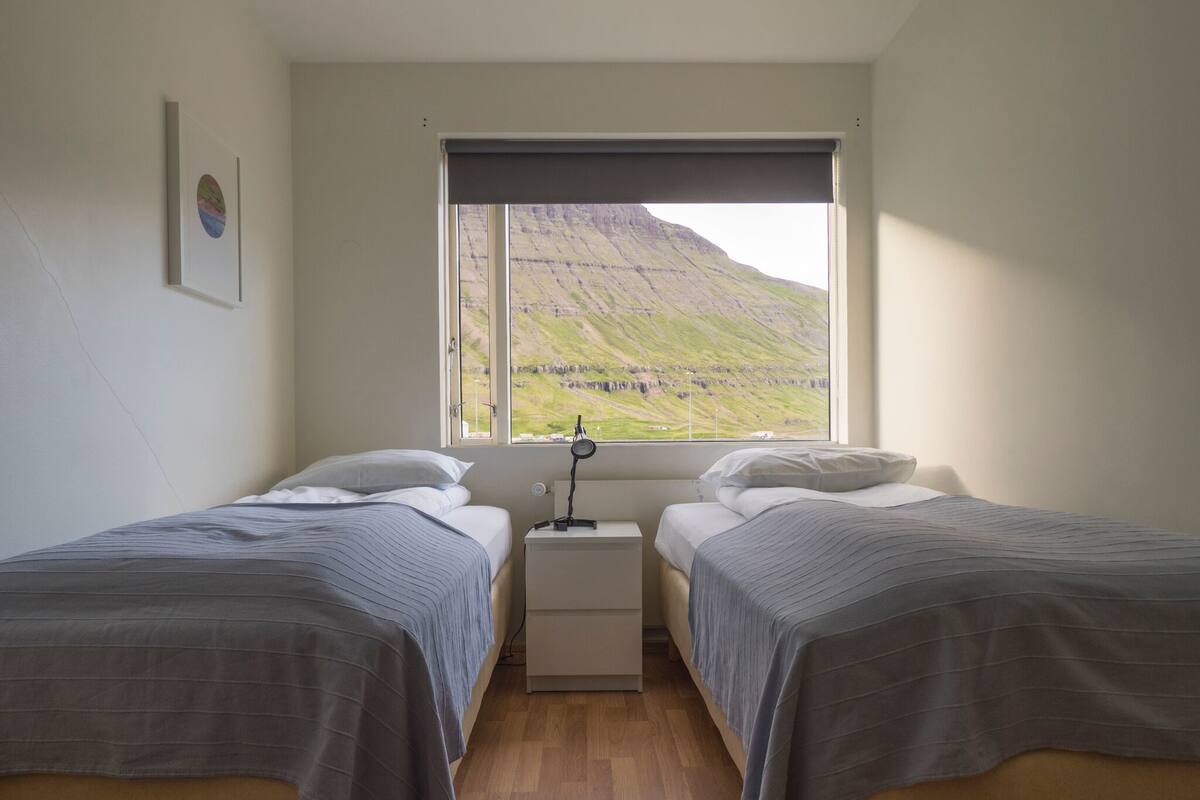 Quarto com duas camas, uma mesa de cabeceira e janela com vista para uma paisagem verde.