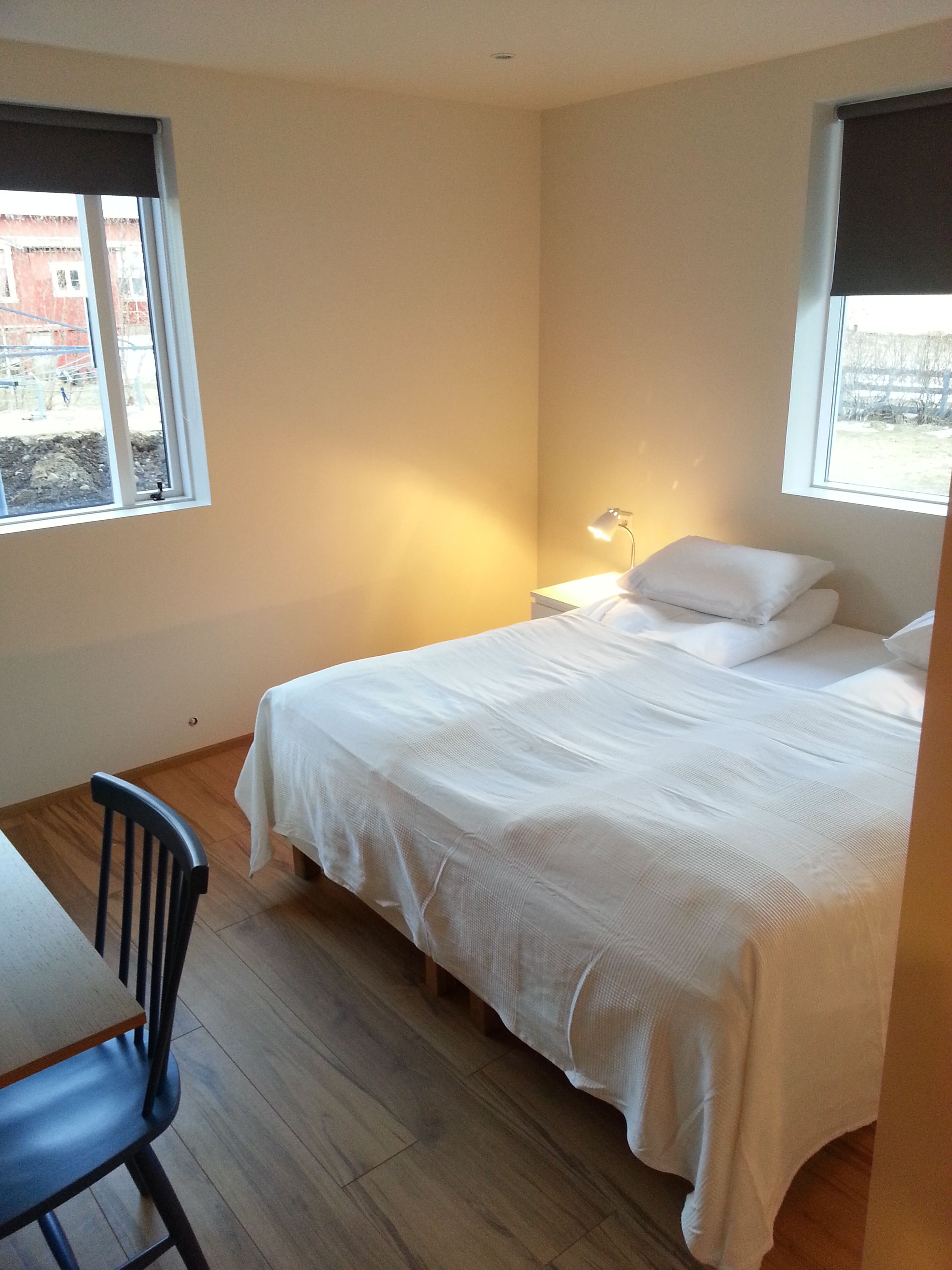 A bedroom with a bed, a desk, and a chair. There are windows on two sides of the room.