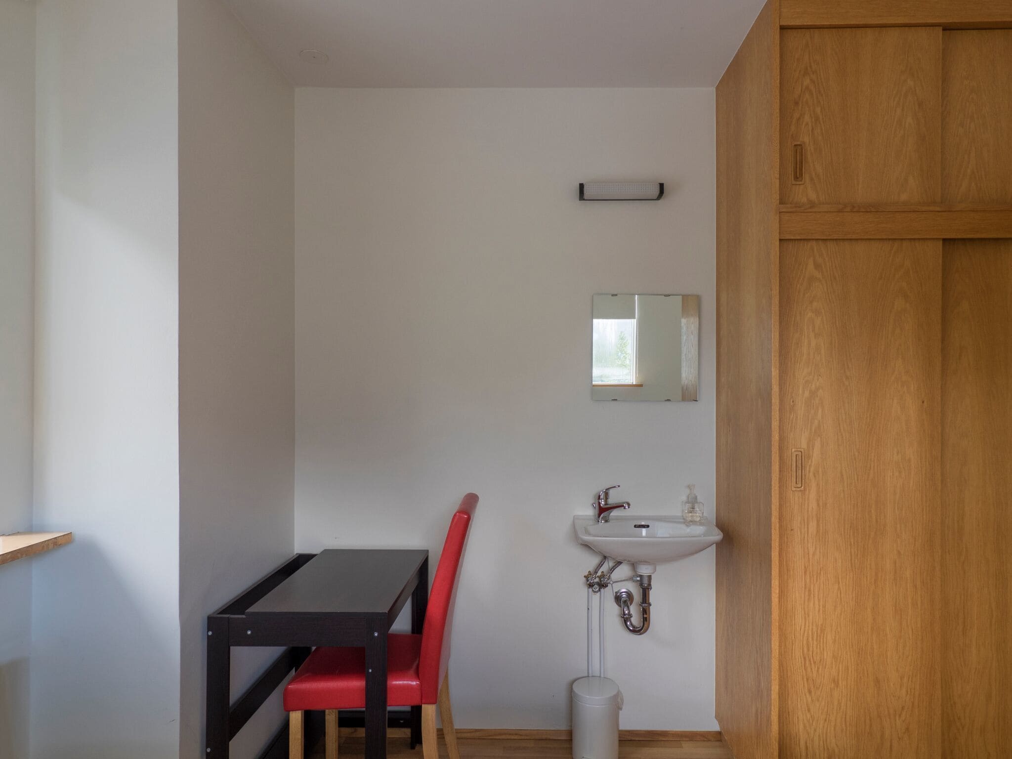 Ein kleiner, kompakter Raum mit einem Holzkasten, einem weißen Waschbecken, einem Spiegel, einem roten Stuhl und einem Tisch.