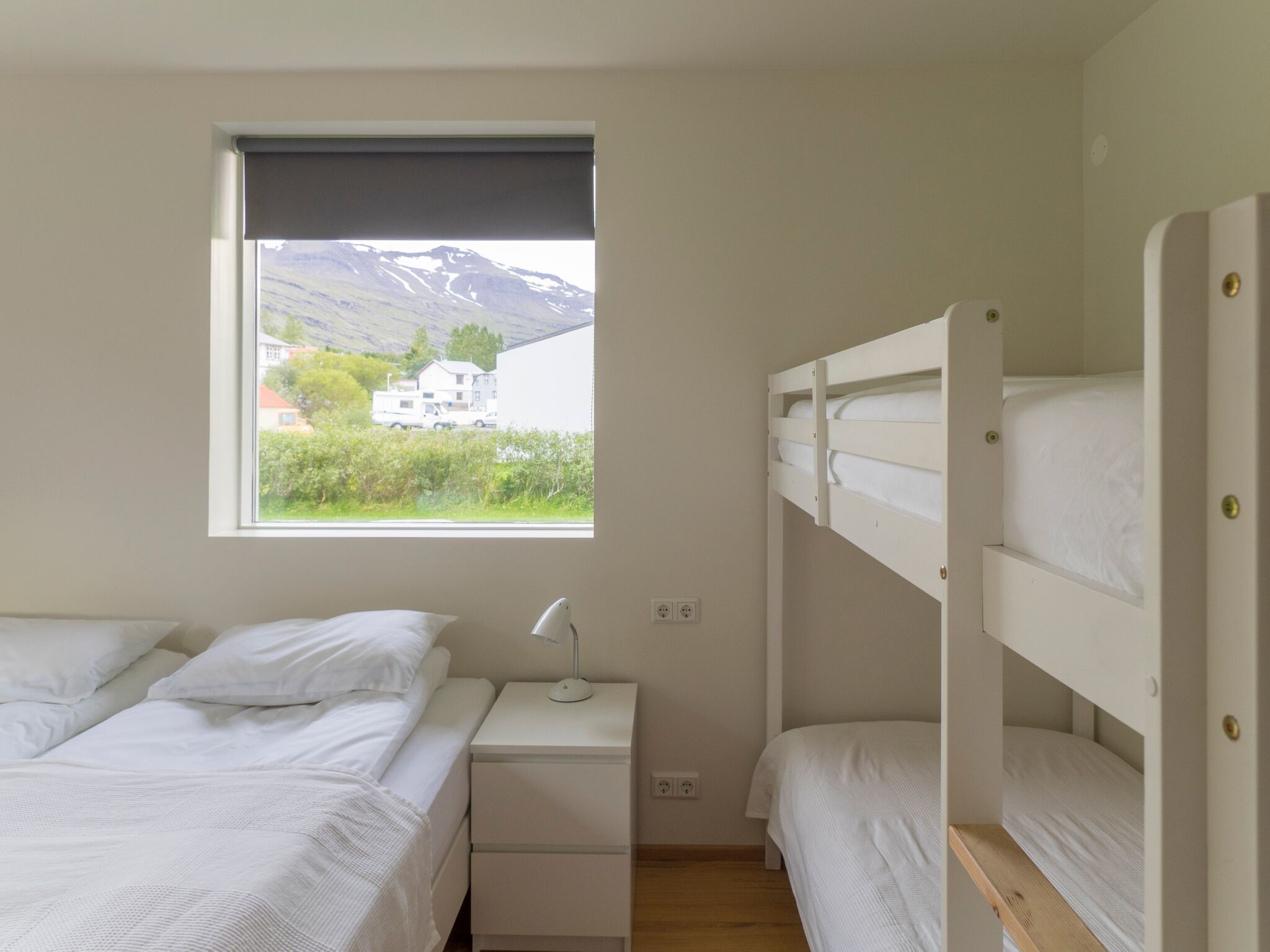 A bedroom with bunk beds, a bedside table with a lamp, and a window with a view of a mountain and buildings.