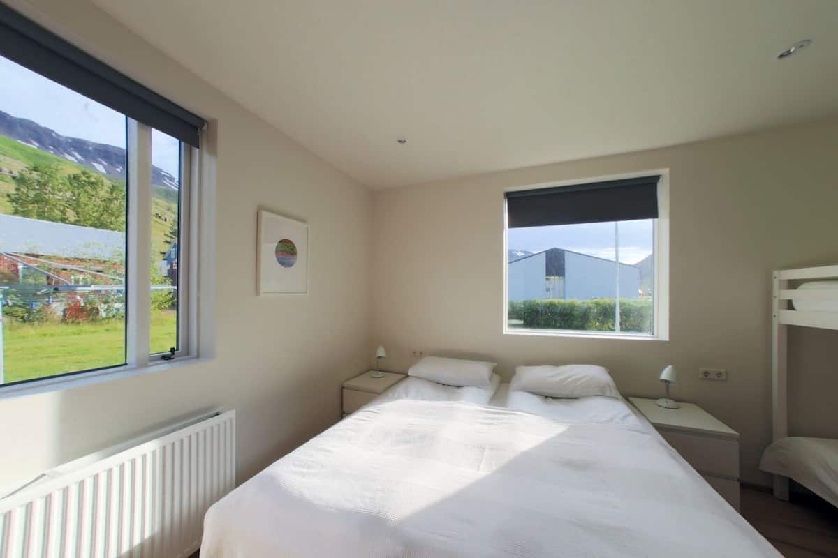 Quarto com uma cama grande, duas mesas de cabeceira, uma janela com vista para áreas verdes e edifícios, e uma cama beliche no canto.