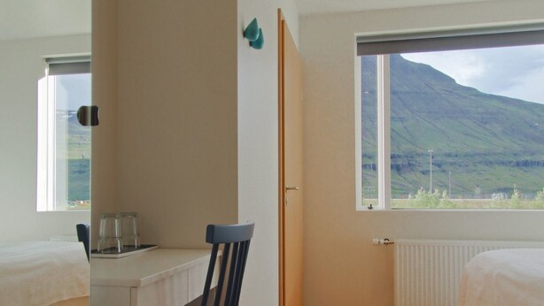 A bedroom with a wooden floor, a desk with a chair, two beds, and a window with a mountain view. - Seyðisfjörður Guesthouse (Seydisfjordur)