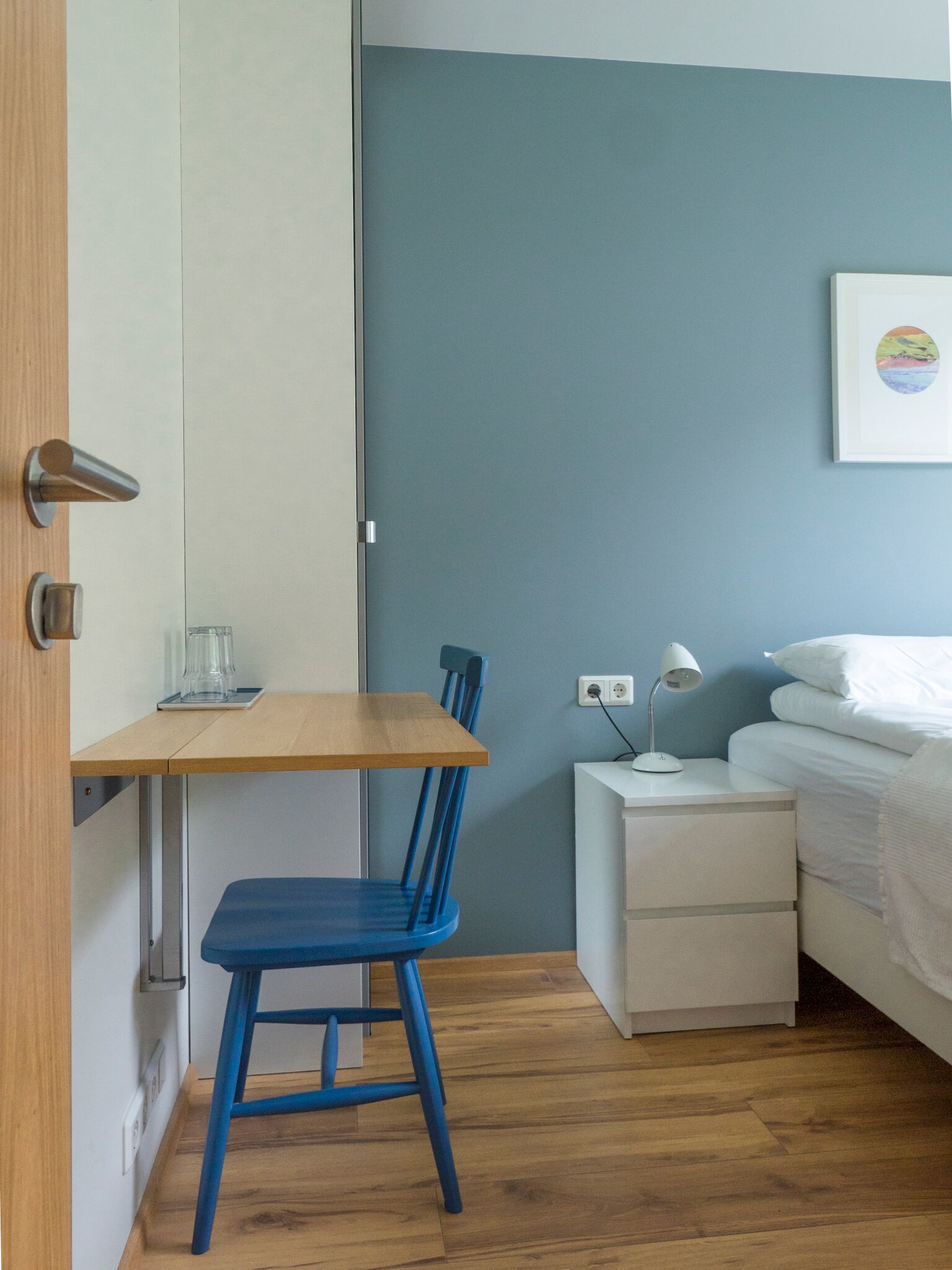 Ein gemütlicher Raum mit Schreibtisch aus Holz, einem blauen Stuhl, einem weißen Nachttisch mit Lampe und einem Bett mit weißer Bettwäsche.
