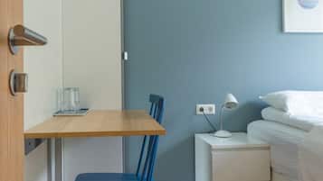 Ein gemütlicher Raum mit Schreibtisch aus Holz, einem blauen Stuhl, einem weißen Nachttisch mit Lampe und einem Bett mit weißer Bettwäsche.