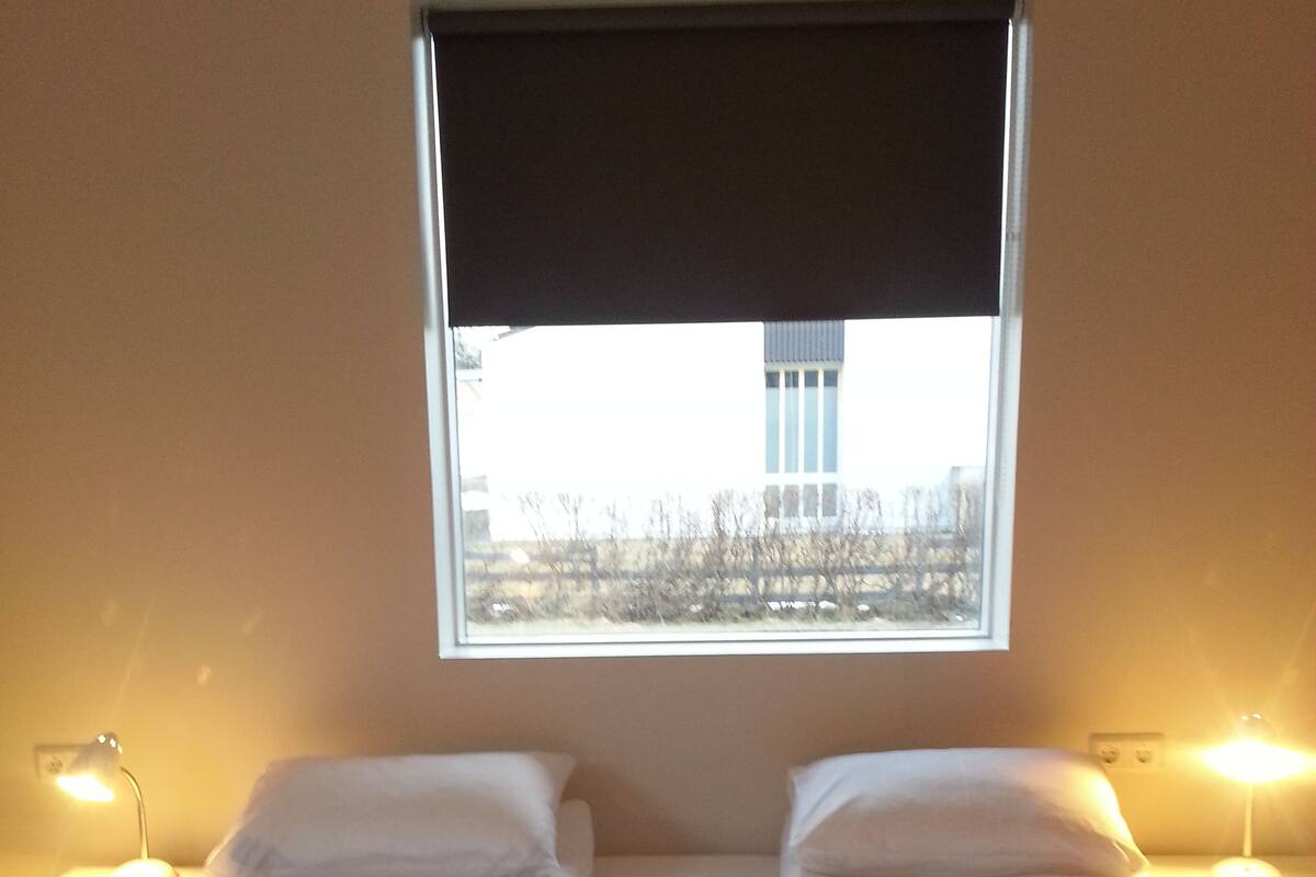 Quarto de hotel com uma cama grande, duas luminárias de cabeceira e uma janela com vista para um edifício.