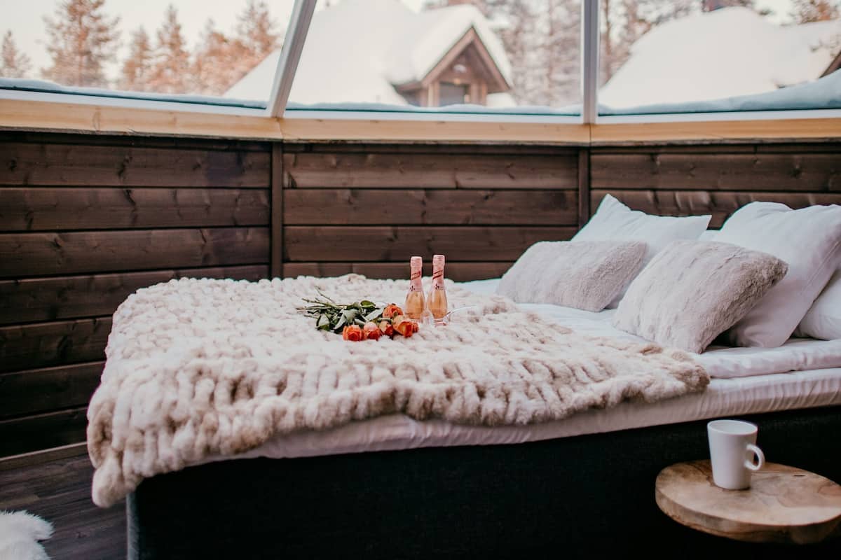 Une pièce chaleureuse avec un lit, une petite table ronde et une fenêtre donnant sur des arbres couverts de neige et un chalet.